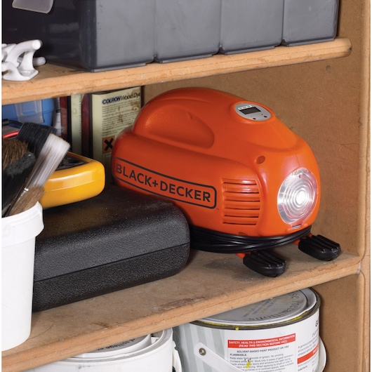 BLACK+DECKER ASI200 orange portable air compressor with coil cord, stored on a wooden shelf among tools and paint cans.