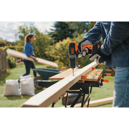 Photo of BLACKDECKER BCD001C1 cordless drill being used on a wooden plank outdoors, with workbench and gloves.