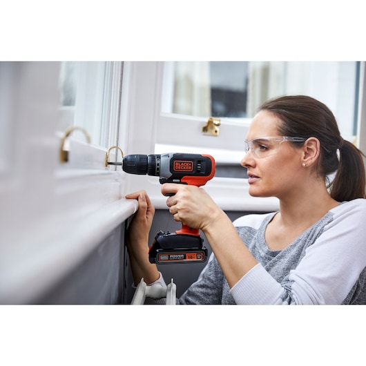 Person using BLACKDECKER BCD001C2 cordless drill on window frame, product visible with branding and battery detail.
