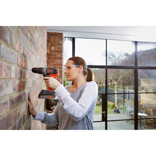 A person using a BLACK+DECKER cordless drill to drill into a brick wall inside a modern home with large windows.
