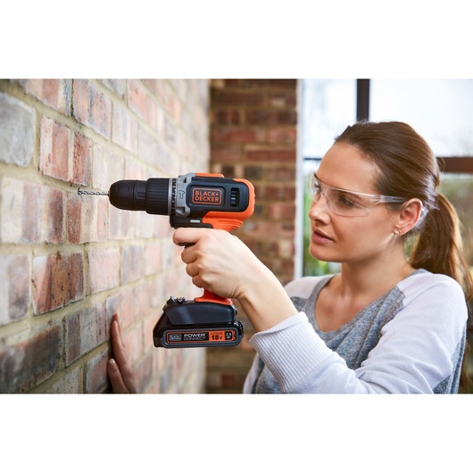 A person using a BLACK+DECKER cordless drill to drill into a brick wall. The individual’s face is blurred for privacy. The drill is orange and black and labeled as an 18V power tool.
