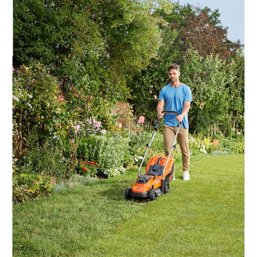 Person mowing lawn with BLACK+DECKER BCMW33185L2 orange cordless lawn mower in a garden.