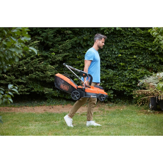 Photo of a man carrying a BLACK+DECKER lawn mower BCMW33185L2 in a garden.