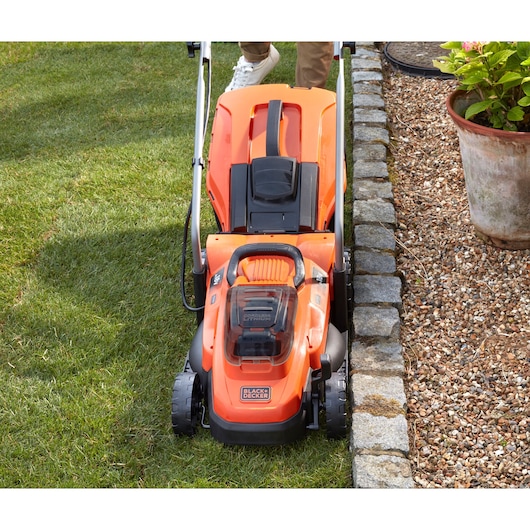 BLACKDECKER BCMW33185L2 cordless orange lawn mower mowing grass next to gravel border, top view.