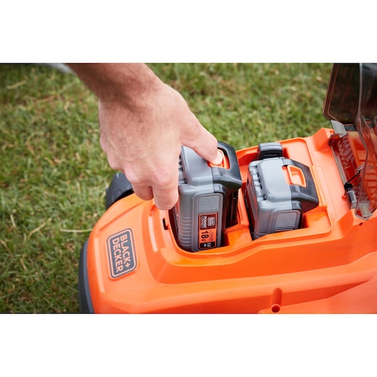 Photo of BLACKDECKER BCMW33185L2 lawn mower with hand inserting battery, orange and gray, outdoor grass background.