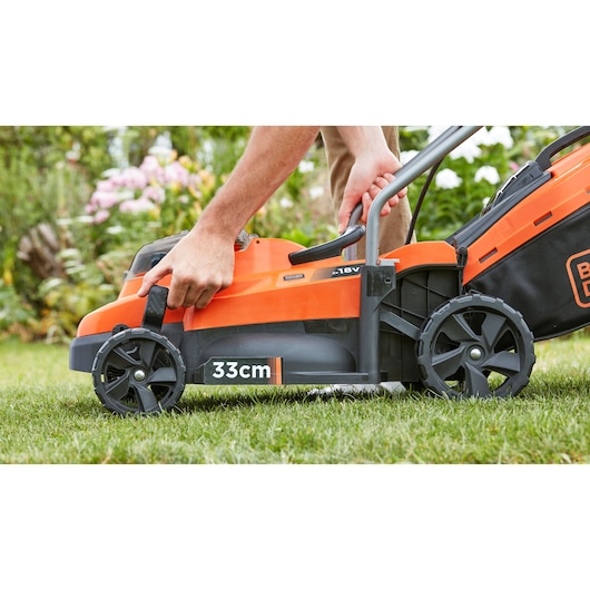 BLACKDECKER BCMW33185L2 lawn mower side view, orange and black, 33cm cutting width, user adjusting handle outdoors.