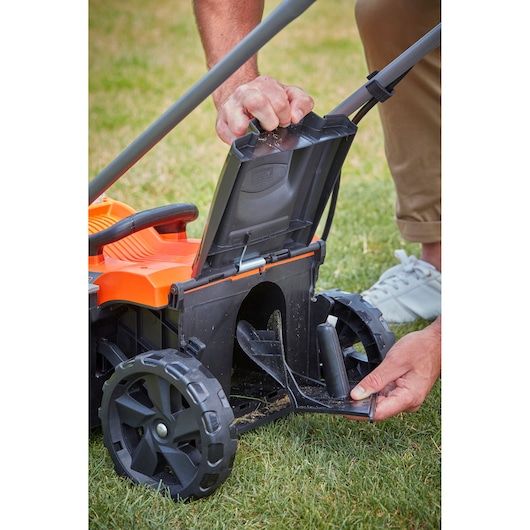 Photo of BLACK+DECKER BCMW33185L2 lawn mower, close-up of rear area with person opening the discharge cover on grass.