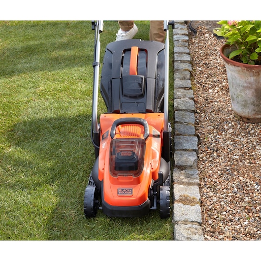 Photo of BLACKDECKER BCMW3336L2 lawn mower in use, orange and black design on grass near stone edging.