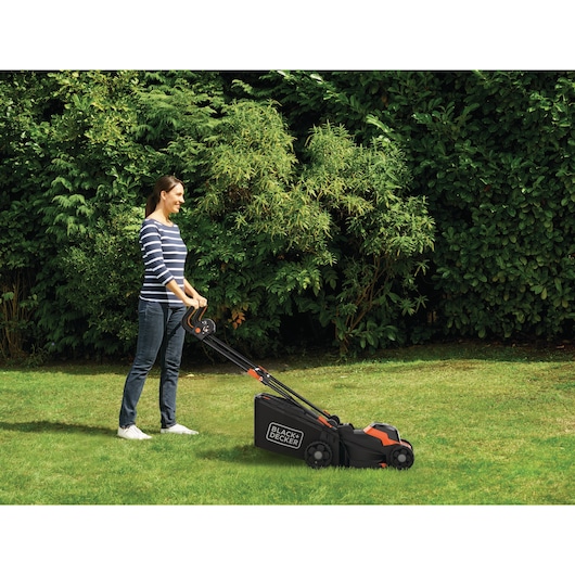 A person is using a BLACK+DECKER lawnmower to cut grass in a garden, with dense green bushes in the background.