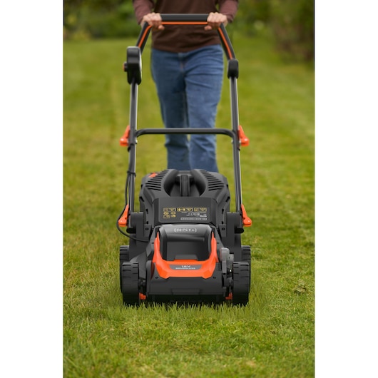 A person is pushing a BLACK+DECKER lawn mower on a green lawn. The image shows the front view of the mower with the person standing behind, holding the handles.