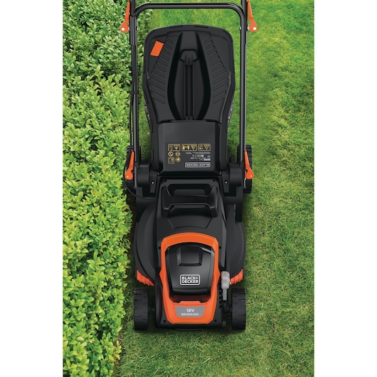 Top view of a BLACK+DECKER 18V cordless lawn mower positioned on a lawn, showing the handle, control panel, and part of a trimmed hedge on the left side.
