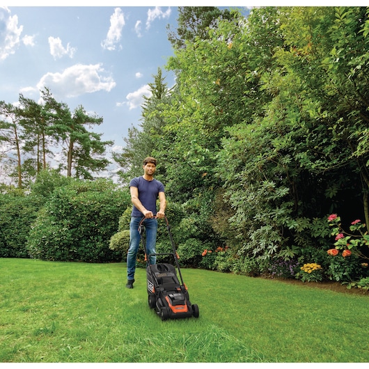 A person with a blurred face is mowing a lawn with a BLACK+DECKER lawn mower in a garden surrounded by green bushes and trees under a blue sky.