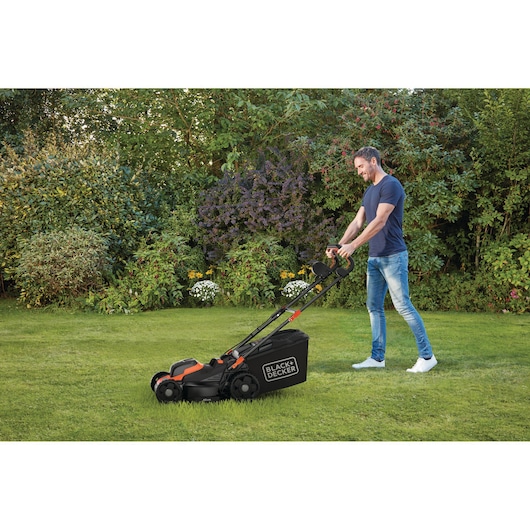 A person is using a BLACK+DECKER lawn mower on a grassy lawn in a garden, surrounded by bushes and flowers.