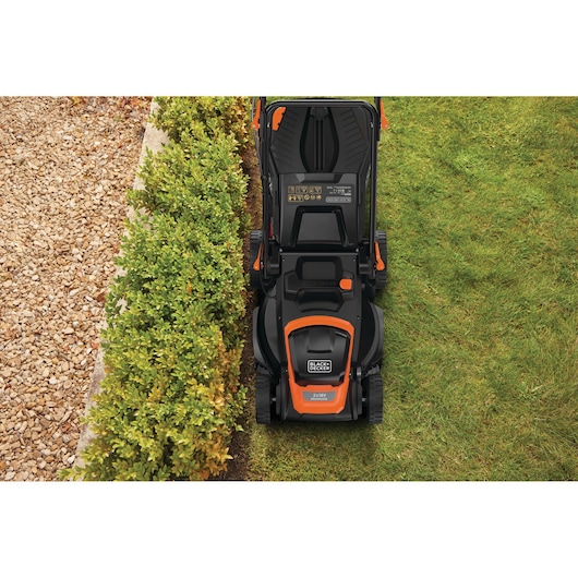Top view of a BLACK+DECKER lawn mower cutting grass near a hedge and a gravel path.