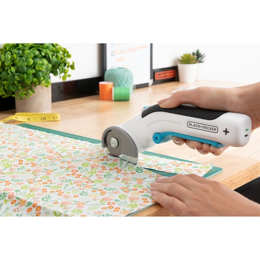 BLACKDECKER BCRC115 rotary fabric cutter, side view, cutting floral patterned fabric on a tabletop.