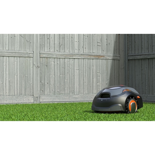 BLACKDECKER BCRMW121 robotic lawn mower on grass in front of a wooden fence, side angle.