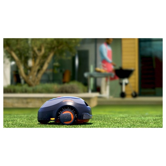 BLACKDECKER BCRMW123 robotic lawn mower on grass, outdoor setting with blurred barbecue in background.