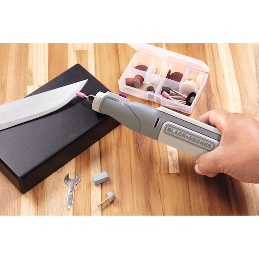 BLACK+DECKER BCRT8IPETK rotary tool sharpening a knife blade, accessory kit and parts visible on wooden table.