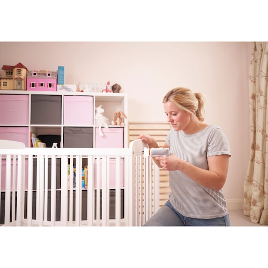 A person is using a BLACK+DECKER tool to assemble or adjust a white baby crib in a nursery. The background features a storage unit with pink and grey boxes, toys, books, and a dollhouse. The person's face is obscured for privacy.