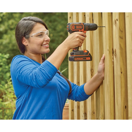 A person in a blue shirt using a BLACK+DECKER 18V lithium cordless drill to drill into a wooden fence outdoors. The person's face is blurred for privacy.