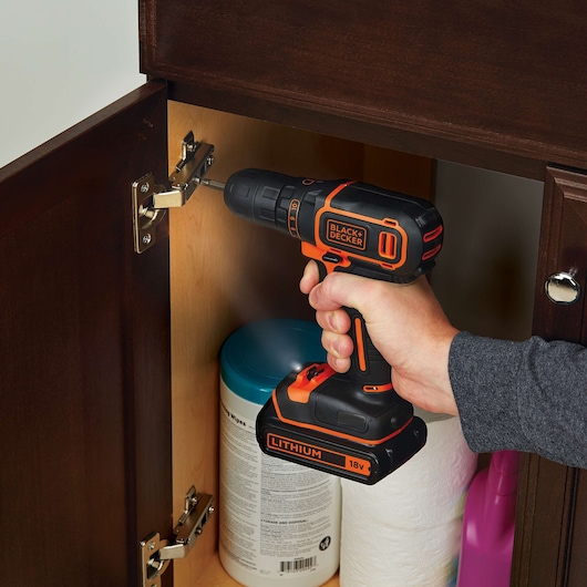 A person is using a BLACK+DECKER 18V lithium cordless drill to screw a hinge inside a wooden cabinet. Cleaning supplies are visible inside the cabinet.