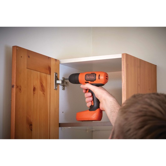 BLACKDECKER BDCD8 cordless drill used to fix cabinet hinge, orange and black body, close-up photo.