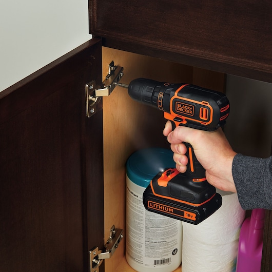 BLACK+DECKER BDCDC18K1B cordless drill used to install cabinet hinge, black and orange design, close-up view.