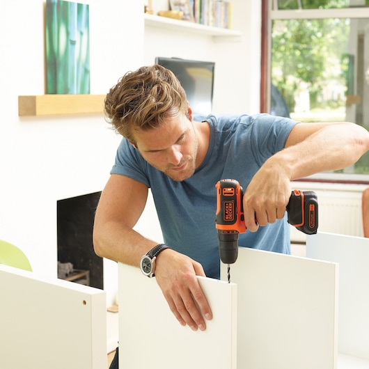 A person using a BLACK+DECKER cordless drill to assemble white wooden panels in a bright indoor setting.