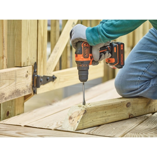 A person wearing gloves uses a BLACK+DECKER cordless drill to drill a hole into a piece of wood outdoors near a wooden fence.
