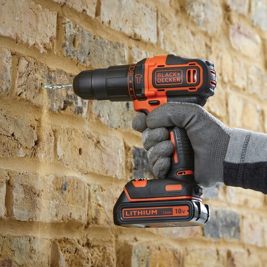 BLACK+DECKER BDCHD18S2KA cordless drill in use drilling into a brick wall, featuring orange and black design.