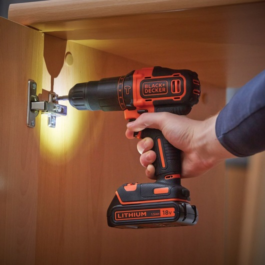 BLACK+DECKER BDCHD18S2KA cordless drill being used on cabinet hinge, orange and black, 18V lithium battery.