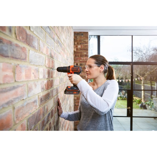 BLACK+DECKER BDCHD18S2KA cordless drill used on brick wall indoors, visible orange and black details.