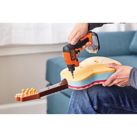A person is using a BLACK+DECKER cordless screwdriver to assemble a colorful toy guitar, with the instrument resting on their lap.