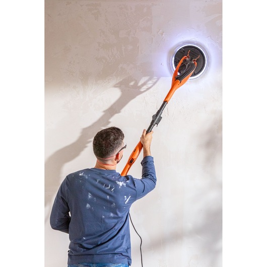 Person sanding a wall with a BLACK+DECKER electric wall sander, bright LED ring around sanding head.