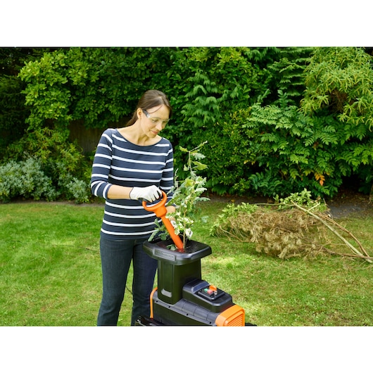 Person using BLACK+DECKER BEGAS5800 garden shredder for branches outdoors, with pile of cut branches nearby.