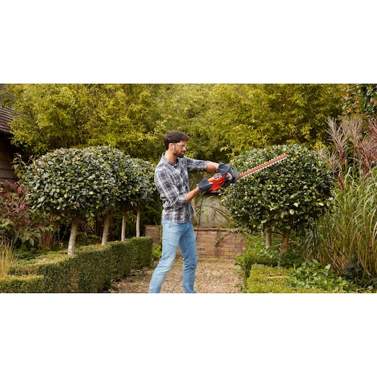 BLACKDECKER BEHT251KIT hedge trimmer used in garden, red and black tool cutting shrubbery outdoors.