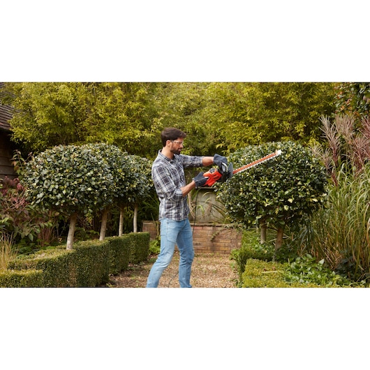 Person trimming bushes with BLACKDECKER BEHTS301 hedge trimmer in a garden.