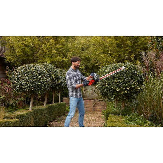 Photography of BLACKDECKER BEHTS501 hedge trimmer in use in a garden trimming green bushes.