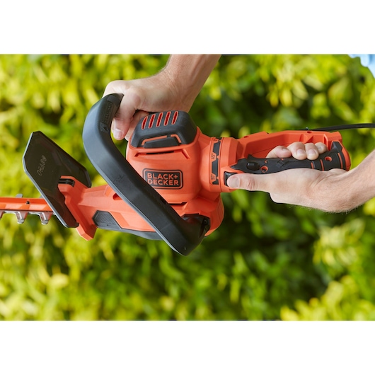 BLACK+DECKER BEHTS551 hedge trimmer held by two hands, orange and black, garden background.