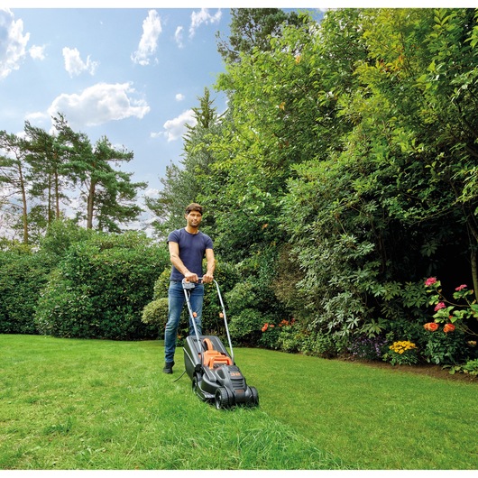 BLACKDECKER BEMW351GL2 lawn mower in use on green grass in a garden, with trees and flowers in background.