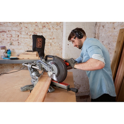 BLACK+DECKER BES700 miter saw cutting wood in workshop, operator wearing gloves and hearing protection.
