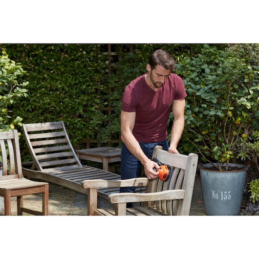 Photo of person sanding wooden outdoor chair with BLACK+DECKER BEW200K detail sander in garden setting.