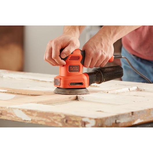 BLACK+DECKER BEW200LSA orange detail sander in use on a wooden surface, hands holding the tool.