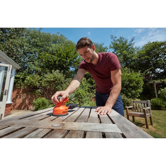 BLACK+DECKER BEW210 random orbital sander used to sand a weathered outdoor wooden table in a garden.