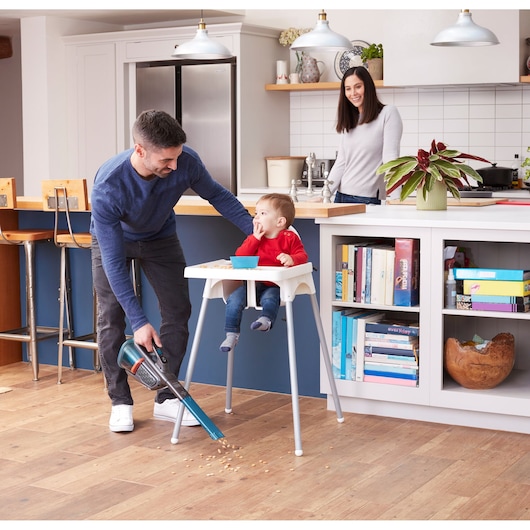 BLACKDECKER BHHV320B handheld vacuum cleaning kitchen floor near high chair with scattered food.
