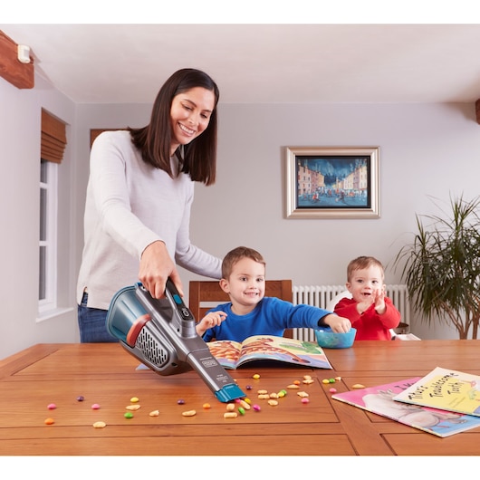 BLACKDECKER BHHV320B handheld vacuum cleaning snack crumbs from wooden table, children sitting nearby.