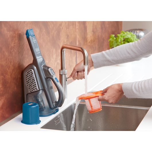 BLACK+DECKER BHHV520BF handheld vacuum filter being washed under a kitchen faucet, with vacuum unit on counter.