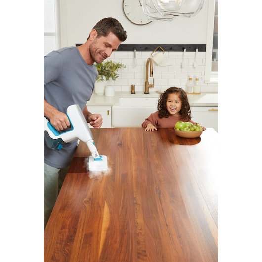 BLACKDECKER BHSM15FX08 handheld steam cleaner used on a wooden kitchen table, bowl of fruit nearby.