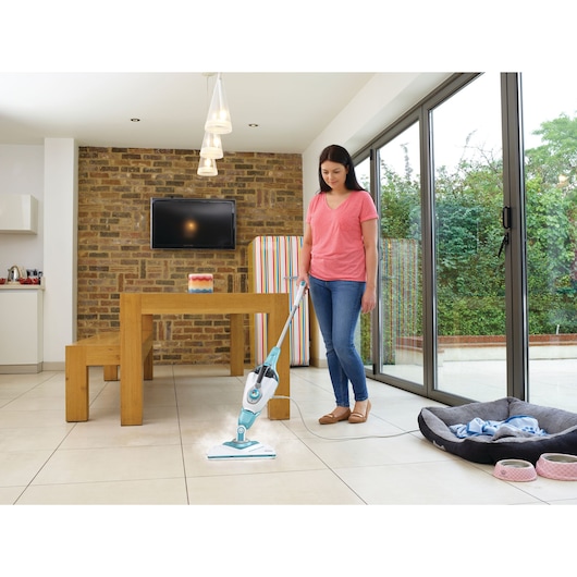 A person uses a BLACK+DECKER steam mop (model BHSM1615DAM) to clean the tiled floor of a modern kitchen and dining area with large glass doors and a wooden table.
