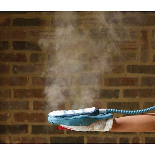 A close-up of a steam cleaning glove being used, emitting visible steam against a brick wall background. The glove is blue and white, attached to a hose, and worn on a person's hand.